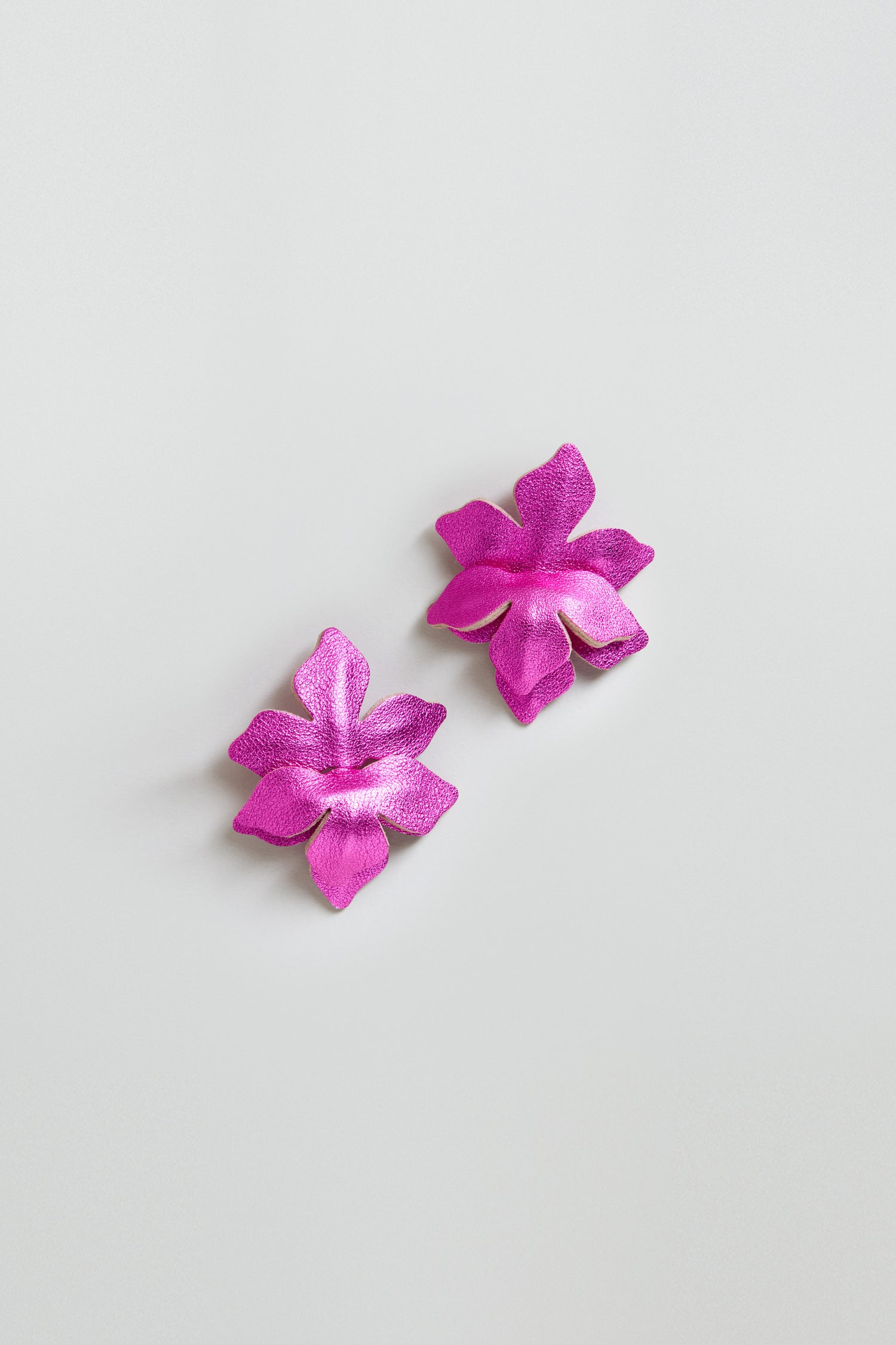 Two vibrant pink metallic leather flower earrings, each with five petals, are displayed side by side on a plain white background, showcasing their handcrafted style and shimmering fuchsia hues.