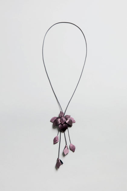The Metallic Leather Flower Necklace features a purple flower pendant and five petal-like charms on a thin metallic leather chain, shown on a white background.
