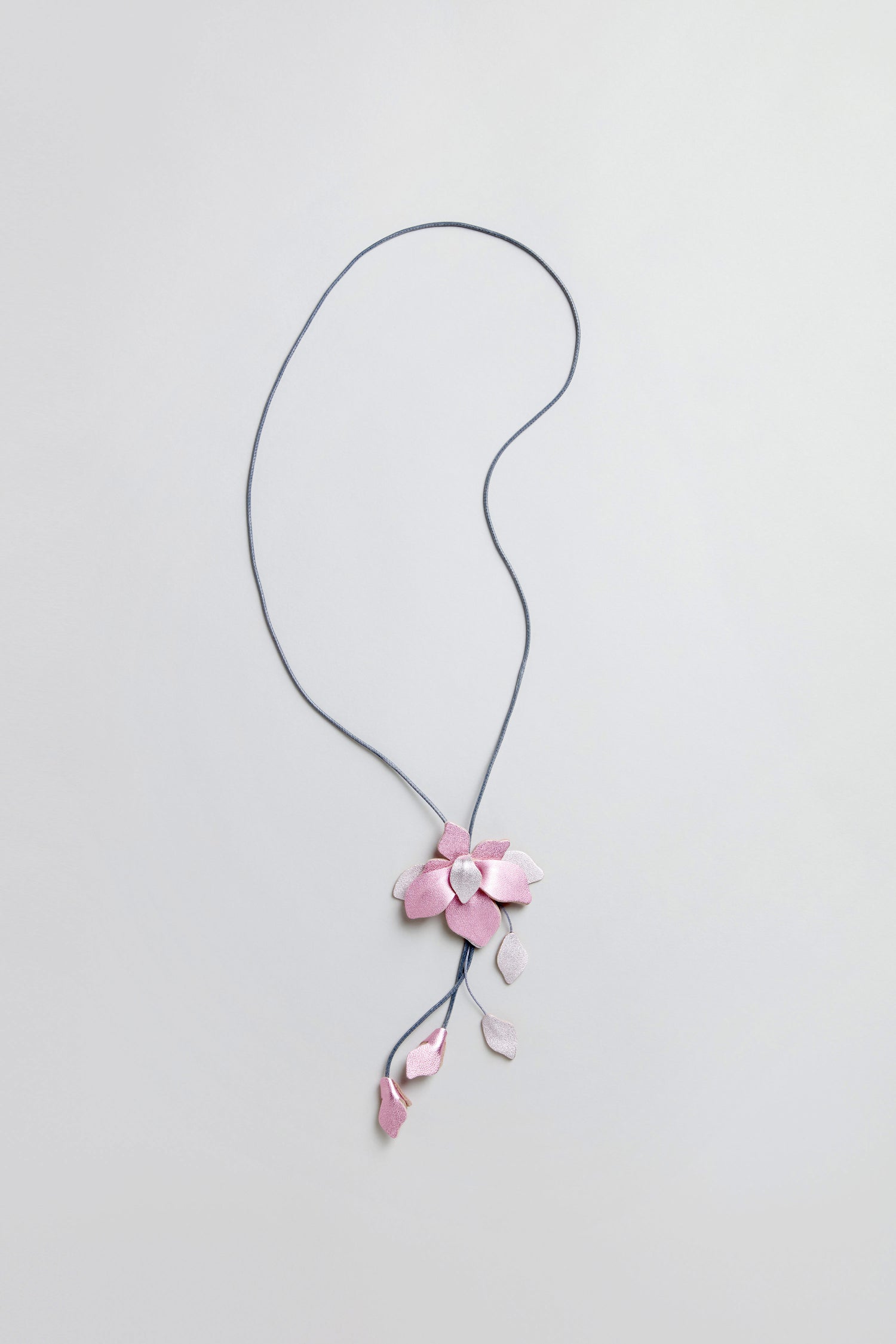The Leather Flower Pendant is a handcrafted necklace with a long blue cord, showcasing a large pink leather flower and small pink leaf charms, all set against a light background.