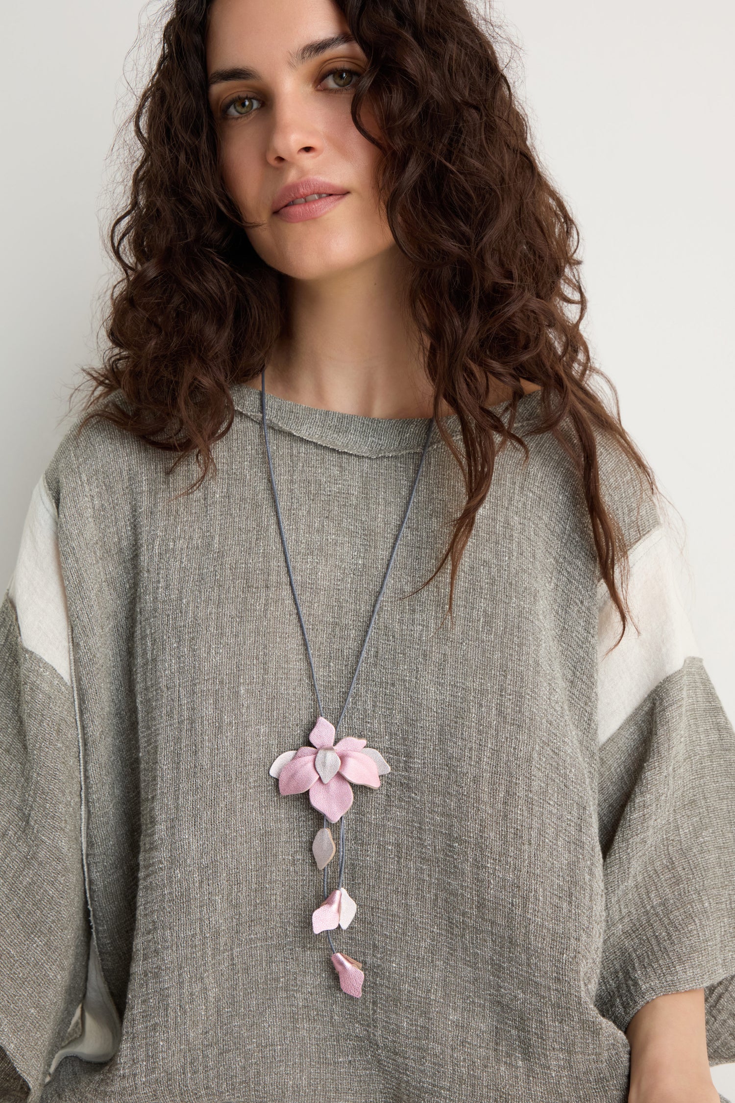 Woman with long curly hair wears a loose gray top and the Leather Flower Pendant necklace featuring a handcrafted pink leather flower, standing against a plain light background.