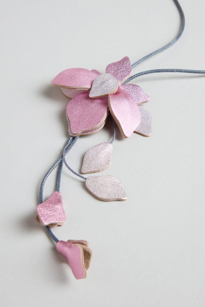 The Leather Flower Pendant is a handcrafted necklace with pink and lilac leather shaped like a blossom with leaves, featuring a long cord and shown against a light grey background.
