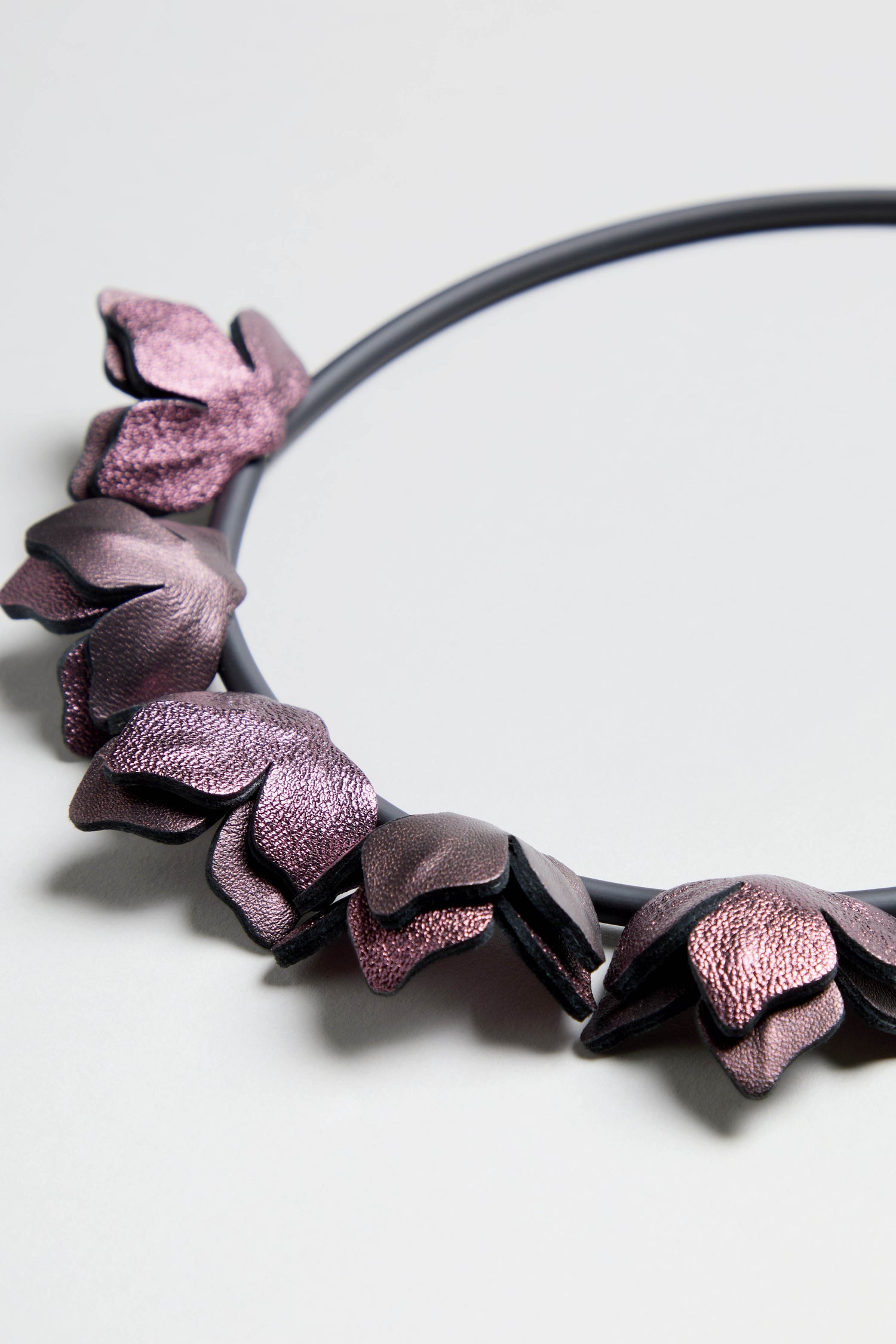 Short Metallic Leather Petals Necklace in black leather features multiple metallic purple, leaf-shaped elements along one side, shown on a light gray background.
