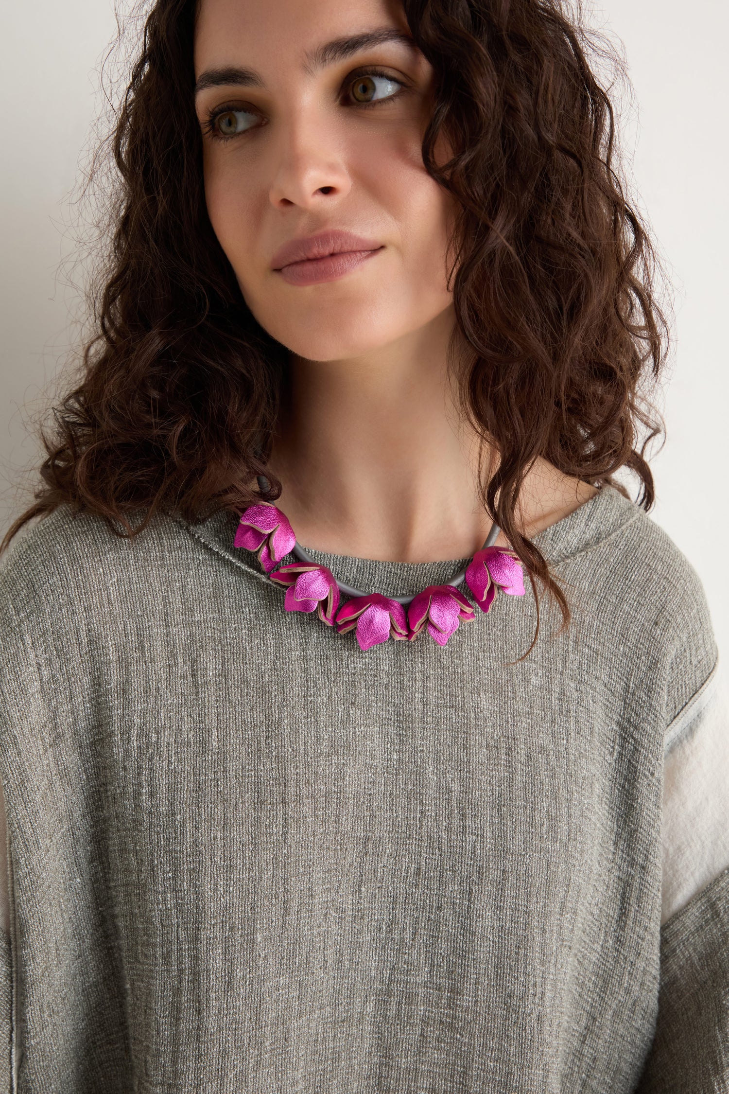 Woman with wavy brown hair wearing a grey textured top and the Short Leather Petals Necklace, which features pink petal shapes, looking slightly to the side.