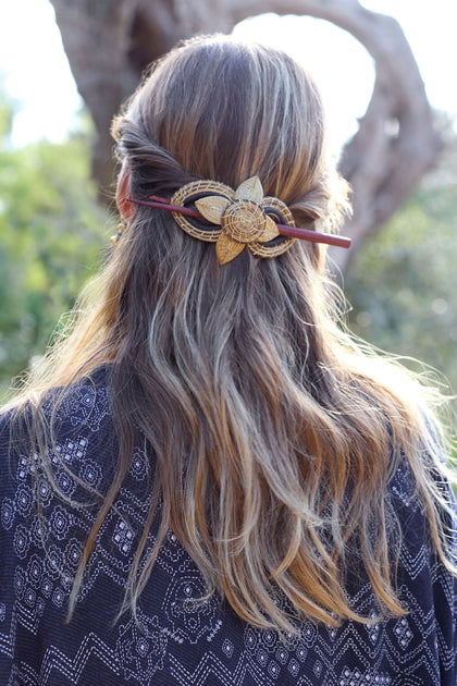 A woman with long hair wears a Golden Grass Woven Flower Hair Pin, showcasing sustainable craftsmanship with a delicate floral design, while outdoor in a patterned garment.