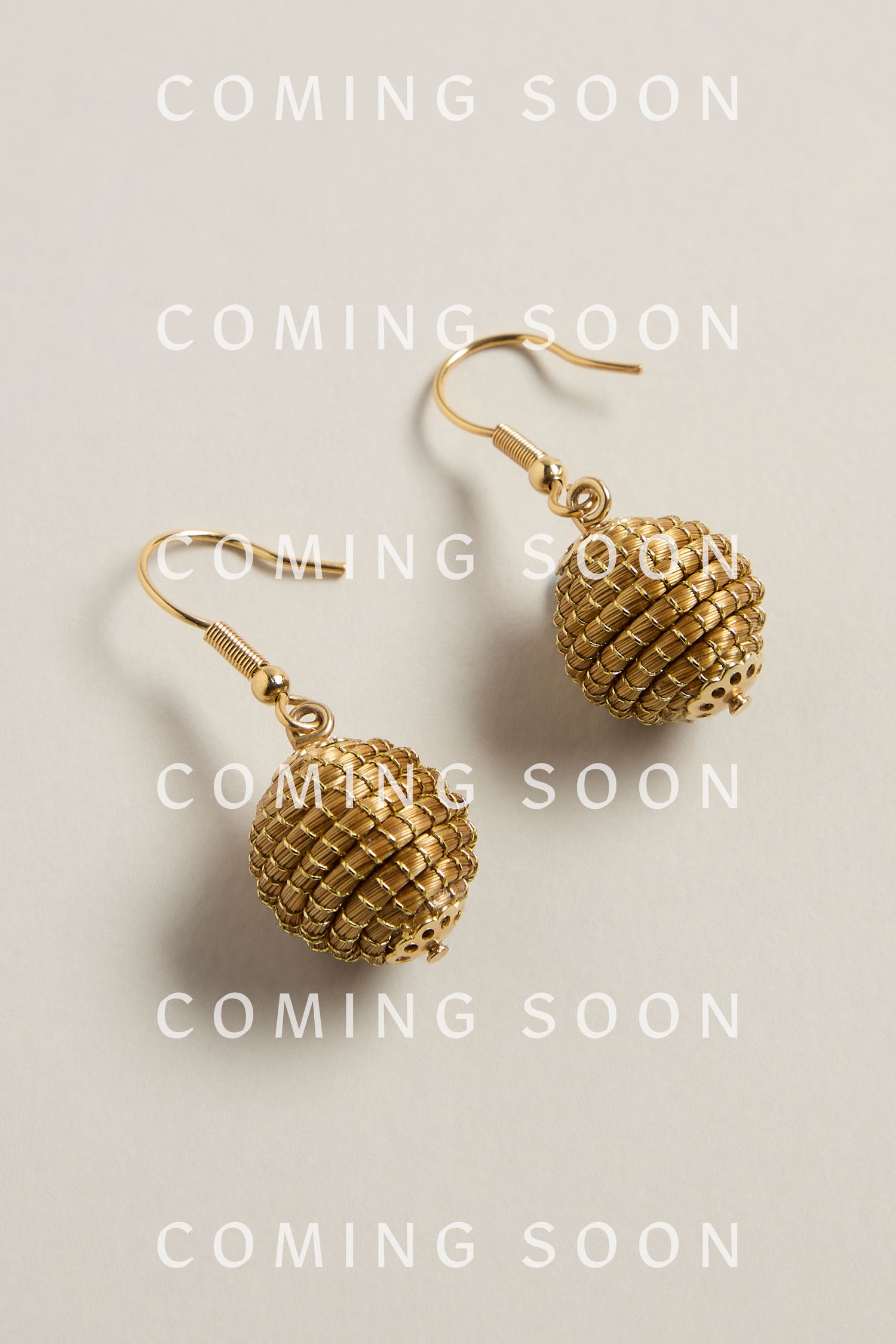 Golden Grass Woven Sphere Earrings glisten on a light background, highlighting the beauty of handcrafted artisan jewelry. "COMING SOON" is overlaid repeatedly, hinting at an exquisite addition crafted with sustainable craftsmanship.