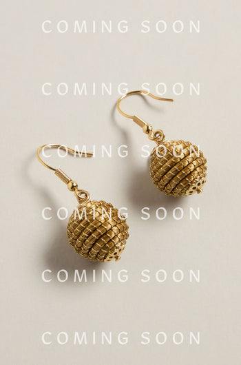 Golden Grass Woven Sphere Earrings glisten on a light background, highlighting the beauty of handcrafted artisan jewelry. "COMING SOON" is overlaid repeatedly, hinting at an exquisite addition crafted with sustainable craftsmanship.