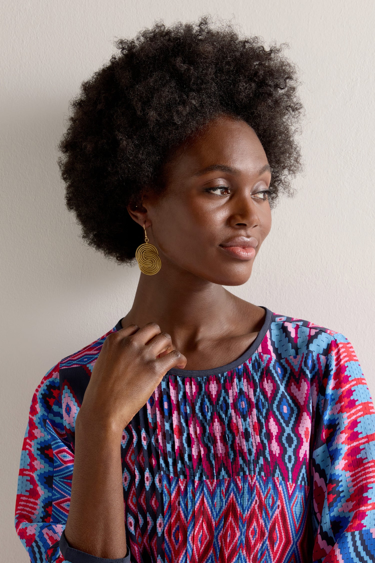 Against a plain backdrop, a woman with an afro hairstyle wears a colorful patterned top and Golden Grass Woven Circles Earrings, gazing to the side.