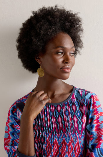 Against a plain backdrop, a woman with an afro hairstyle wears a colorful patterned top and Golden Grass Woven Circles Earrings, gazing to the side.