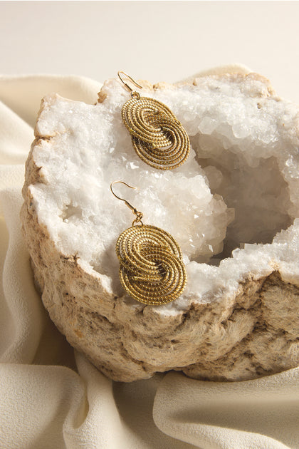 The Golden Grass Woven Circles Earrings, showcased on a textured white geode against a soft fabric backdrop, exemplify handcrafted jewelry with their sustainable design blending elegance and environmental consciousness.