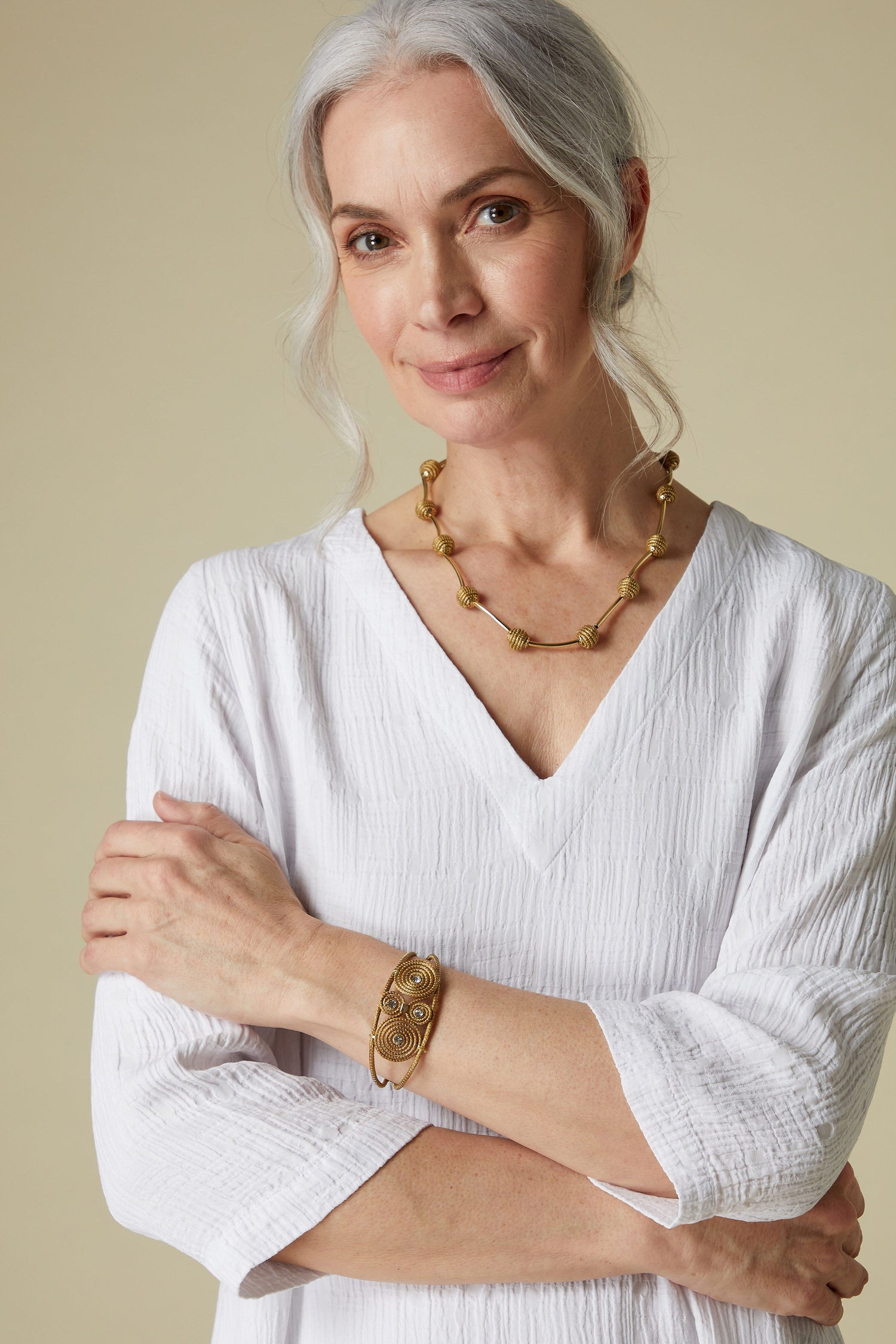 A mature woman with gray hair, wearing a white blouse and gold jewelry, including a Golden Glass Spiral Rhinestone Bracelet, smiling gently at the camera.