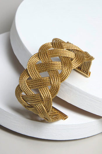 The Golden Grass Woven Bracelet, a testament to exquisite artisanal craftsmanship, is elegantly showcased on a sleek white cylindrical surface.