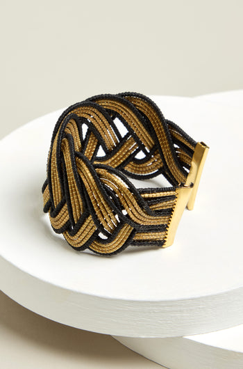 The Gold and Black Grass Woven Bracelet, showcasing a sustainable design, elegantly rests on a white round surface.