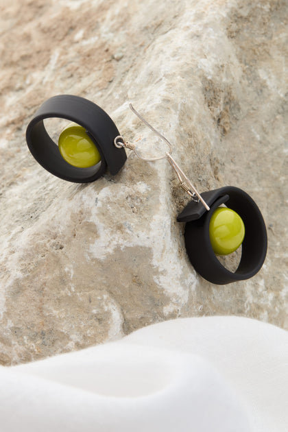 The Tempo Earrings are a pair of black earrings featuring circular designs and adorned with green spherical beads, crafted from durable materials and displayed on a textured stone surface.