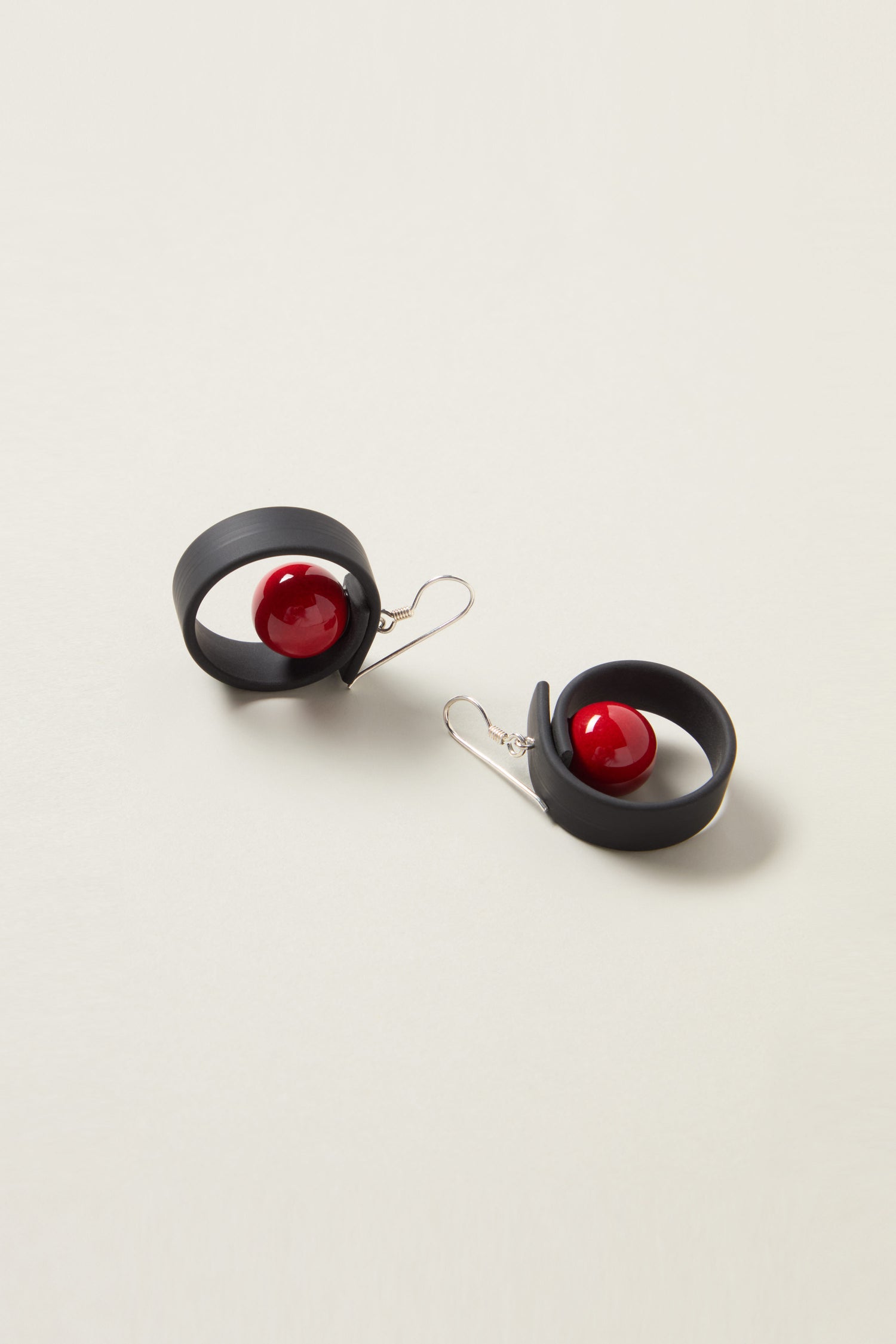 The Tempo Earrings are a pair of modernist French jewelry pieces in black with a spiral design, featuring a single red spherical bead at the center. They are crafted from durable materials and elegantly placed on a light-colored surface.