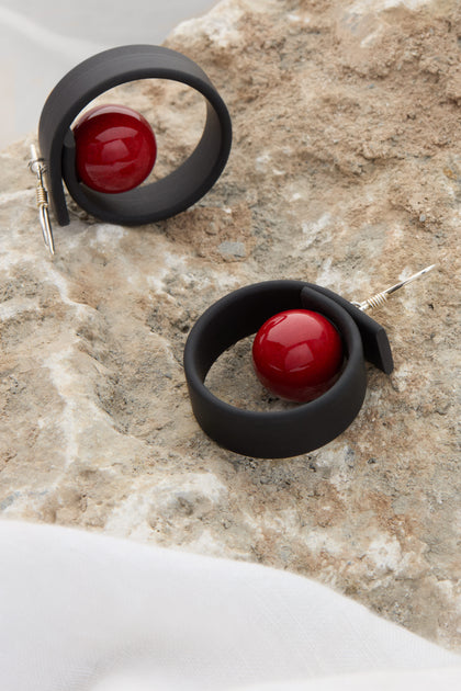 Two Tempo Earrings, featuring black coils with red spherical beads, crafted from durable materials and inspired by modernist French jewellery design, placed on a textured stone surface.