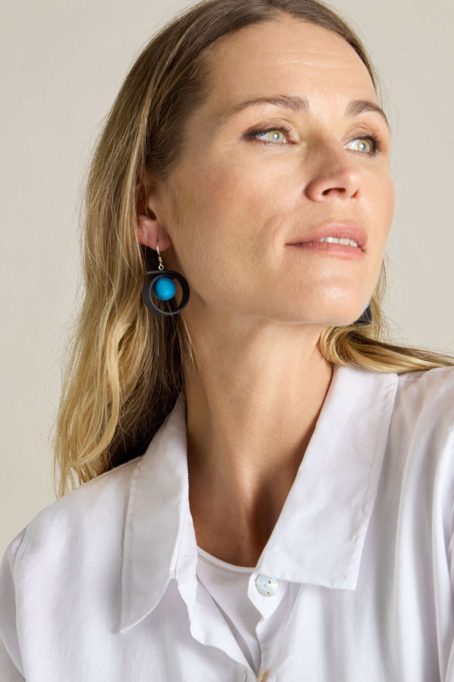 A woman with long blonde hair wearing Tempo Earrings, a piece of modernist French jewellery, and a white shirt looks upwards.