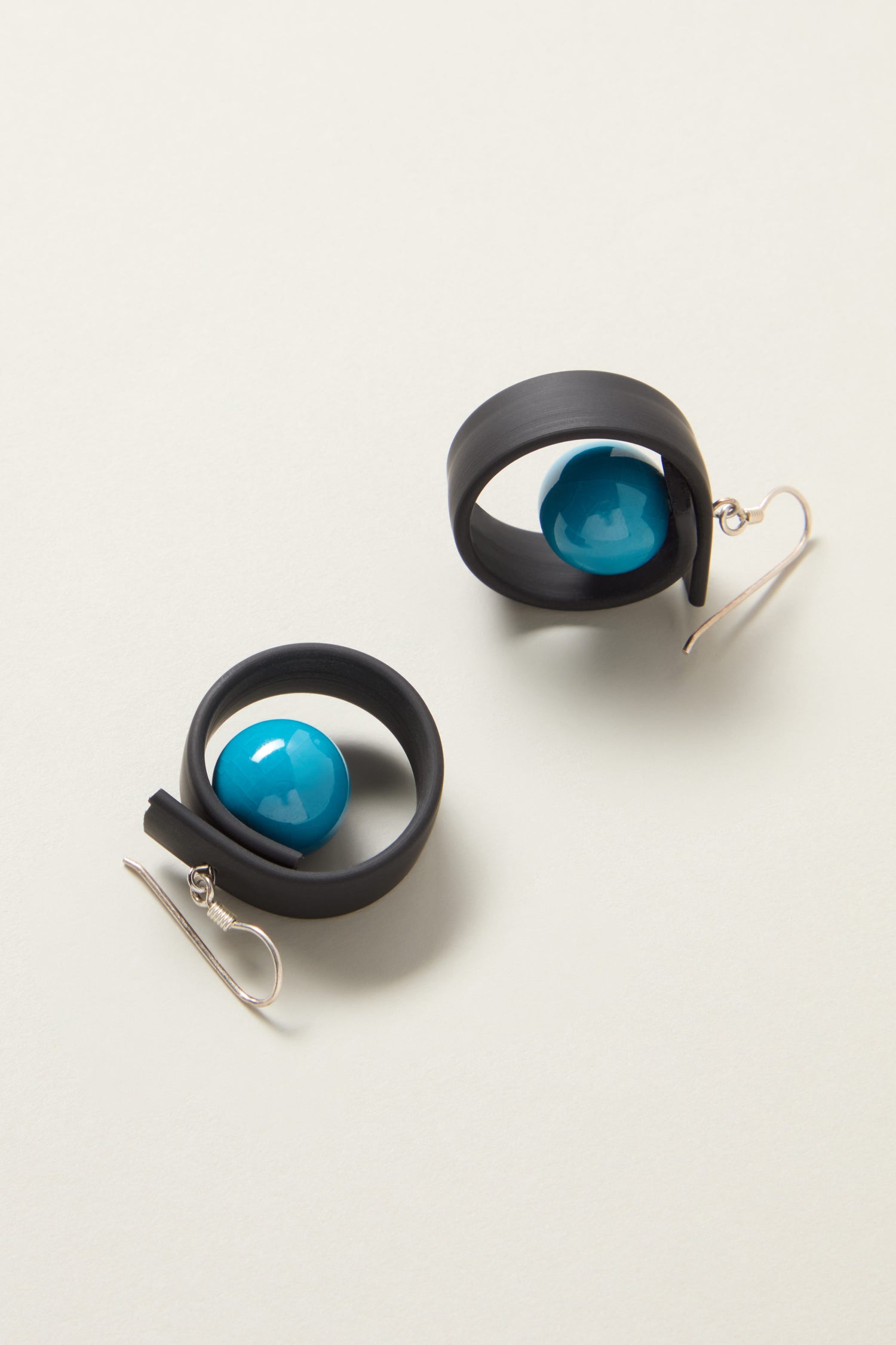 The Tempo Earrings showcase black loops embracing a single blue bead, complemented by silver hooks for wearing. Made from durable materials, these earrings capture modernist French jewelry charm while offering enduring elegance.