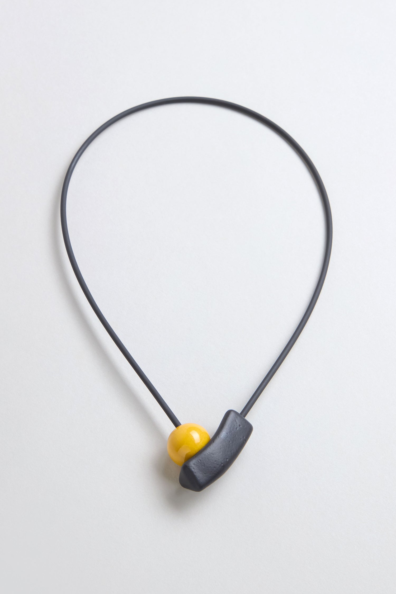The Atom Necklace is a minimalist ceramic accessory with a thin black cord, featuring geometric elements—a yellow bead and curved black piece—displayed against a light background.