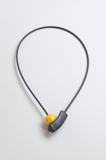 The Atom Necklace is a minimalist ceramic accessory with a thin black cord, featuring geometric elements—a yellow bead and curved black piece—displayed against a light background.