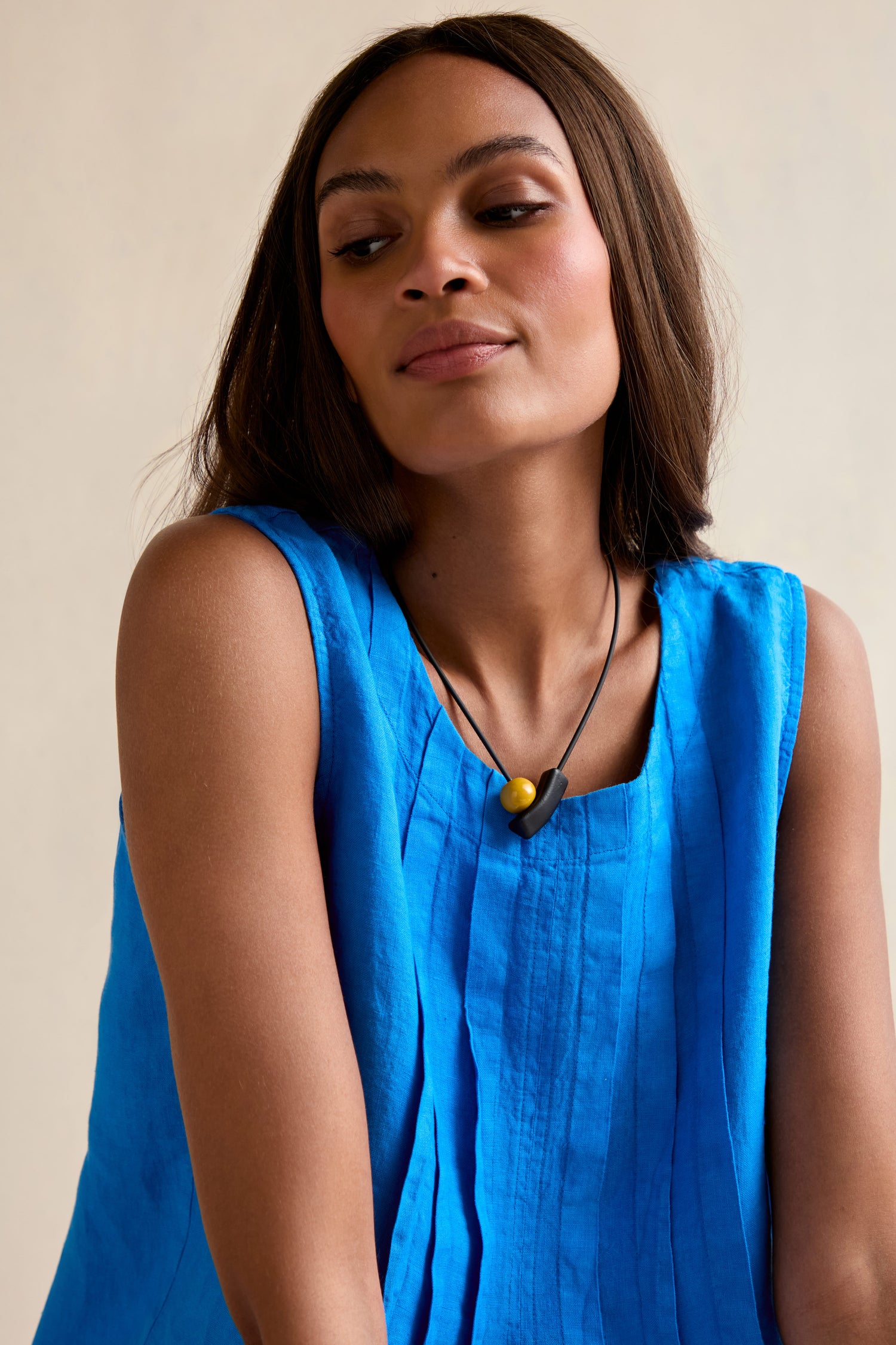 A woman with straight brown hair wears a sleeveless bright blue top and the Atom Necklace—featuring black geometric elements with yellow and black beads—looking down and to the side against a neutral background.