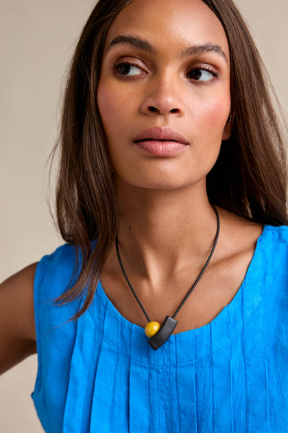 A woman in a sleeveless blue top wears the Atom Necklace, featuring geometric black and yellow enamel beads, as she looks to the side against a neutral background.