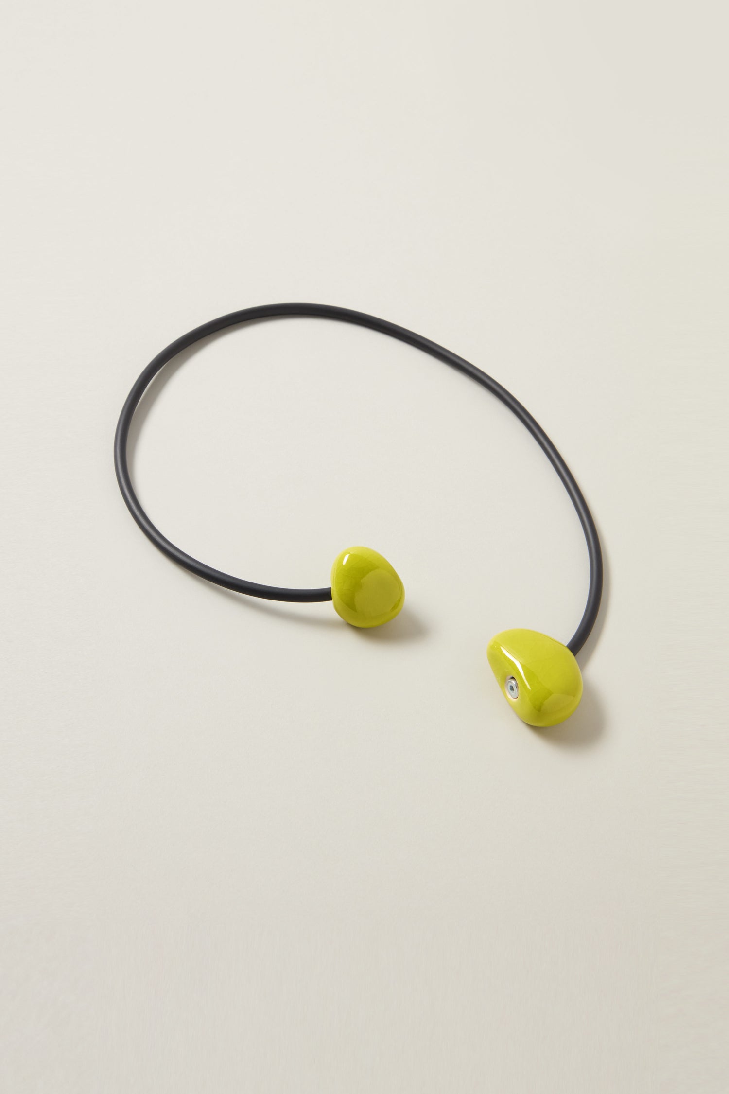 The Short Ceramic Pebble Necklace, featuring a black cord adorned with two green, bean-shaped ceramic ends, exemplifies contemporary style as it lays on a white surface. The necklace includes a magnetic clasp for easy wear.