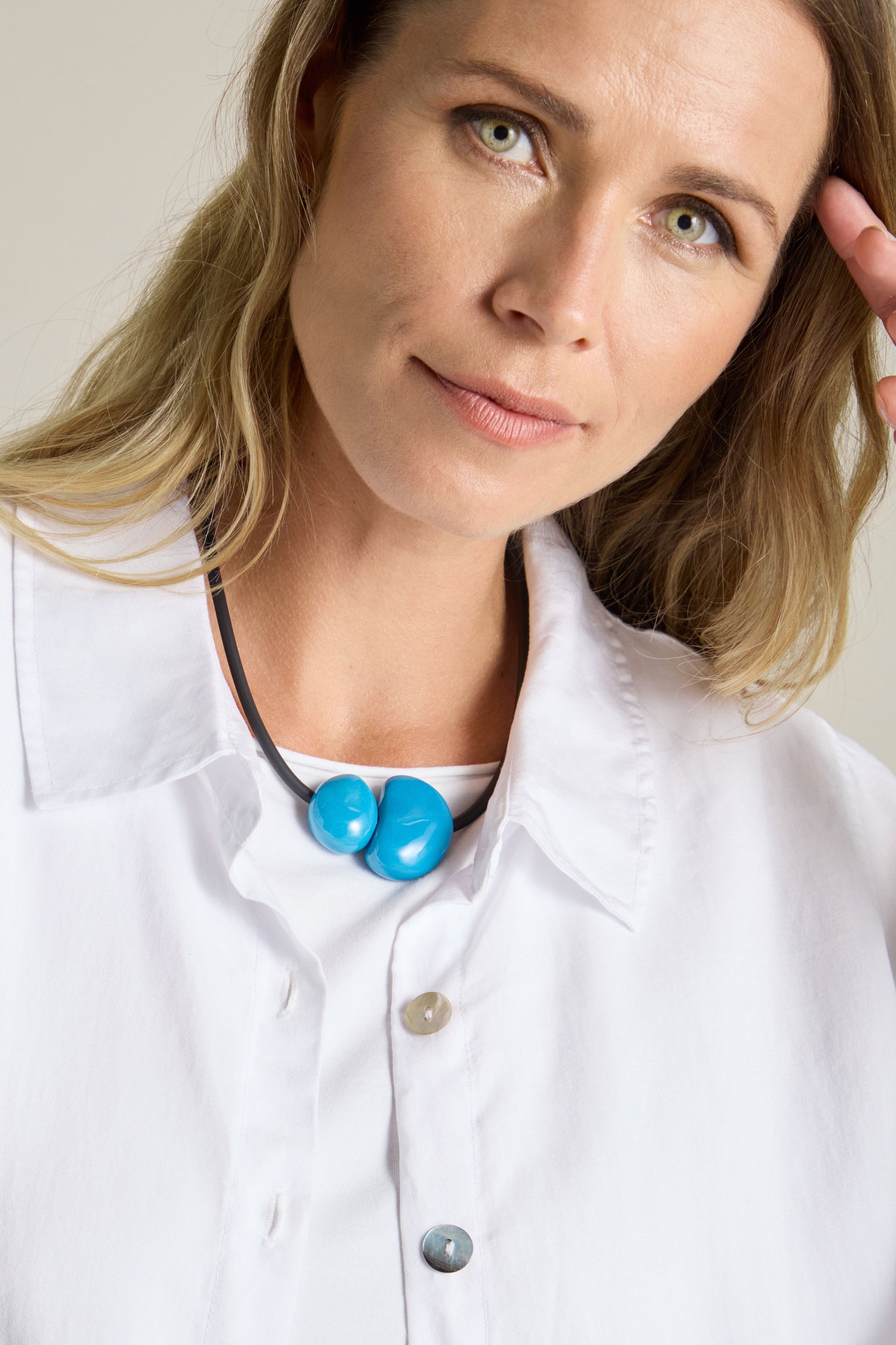 A person with light hair, wearing a white shirt and a Short Ceramic Pebble Necklace featuring two blue beads, places their hand near their head while looking slightly off-camera.