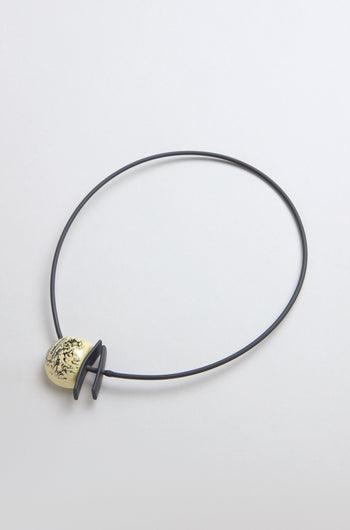 The Calligraphy Necklace features a minimalist design with a thin black cord and a gold ceramic pendant embellished with an abstract black pattern.