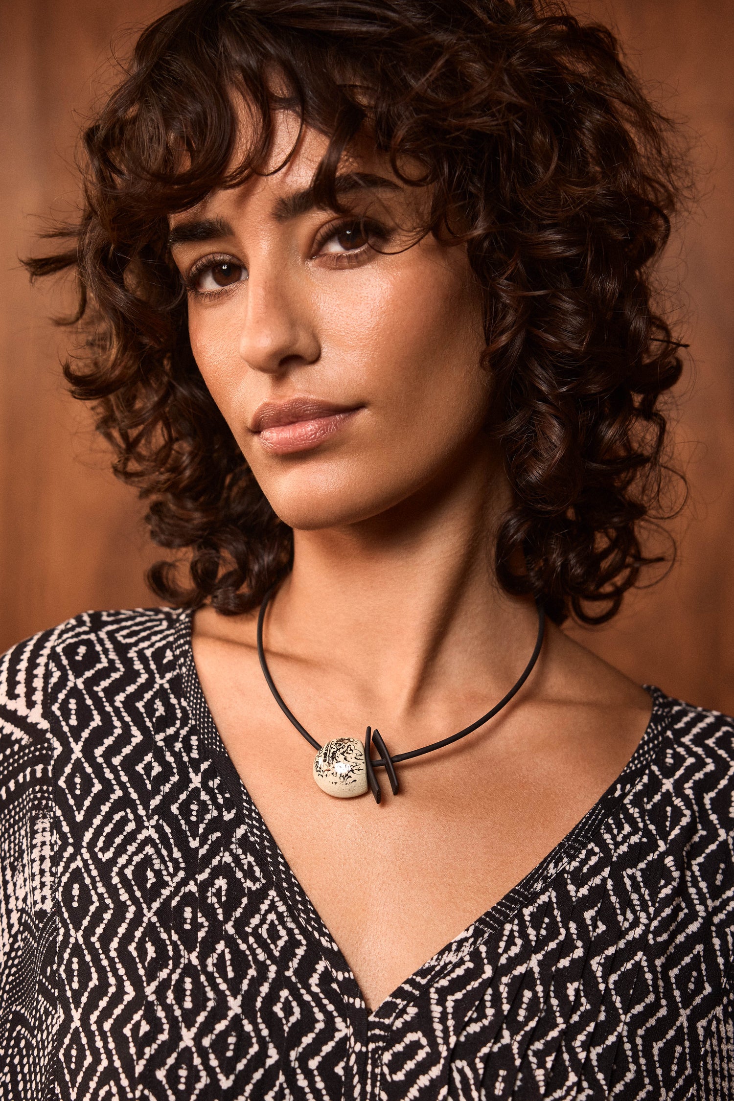 A woman with short curly brown hair wears a black and white patterned top and the handmade Calligraphy Necklace featuring a round ceramic pendant, standing in front of a wooden background.