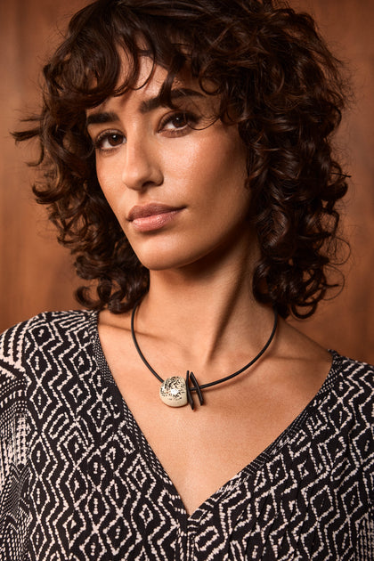 A woman with short curly brown hair wears a black and white patterned top and the handmade Calligraphy Necklace featuring a round ceramic pendant, standing in front of a wooden background.