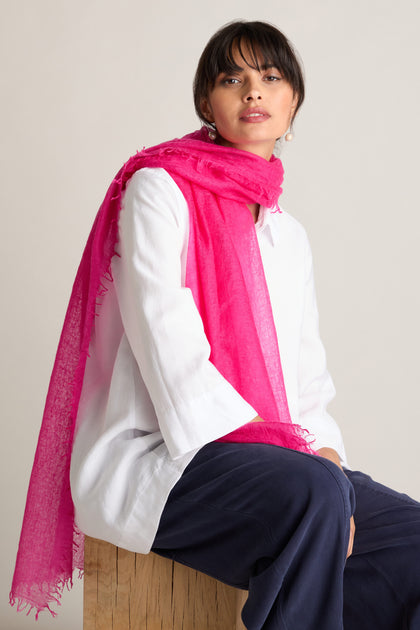 A person wearing a white top, black pants, and a Soft Weave Cashmere Scarf with a raw-edge tassel finish sits on a wooden stool, facing slightly to the right with a neutral expression.