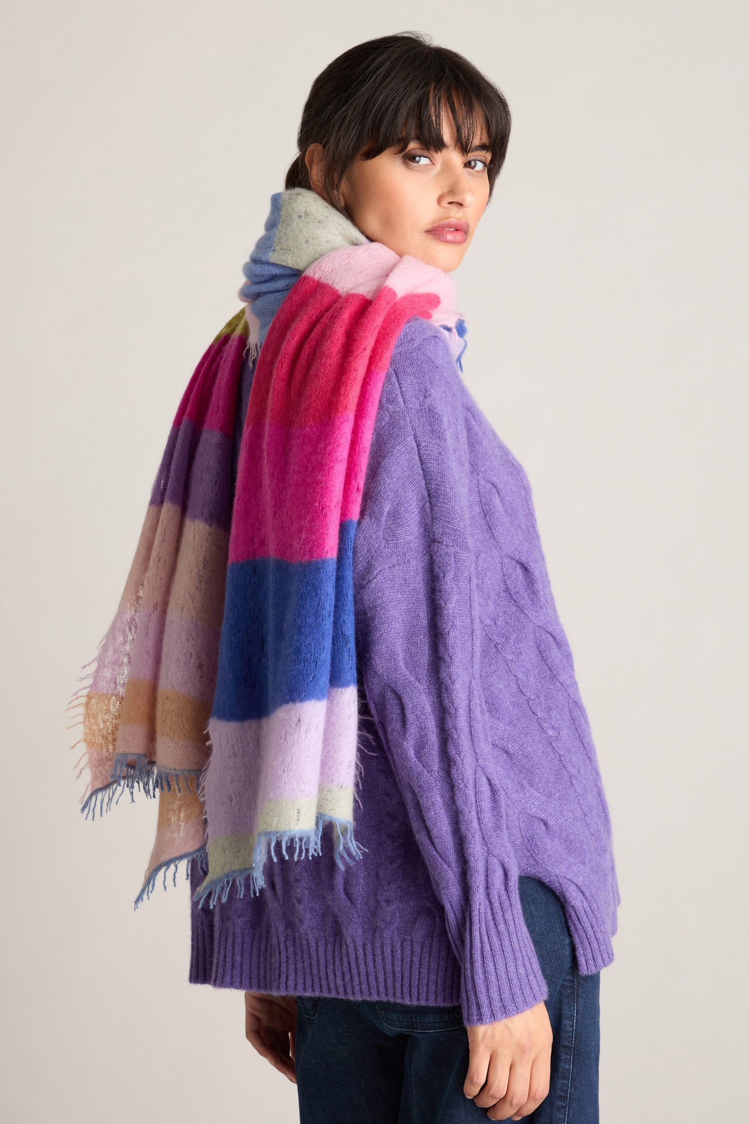 A person with dark hair wears a purple sweater and the Rainbow Stripe Cashmere Scarf, looking over their shoulder against a plain background.