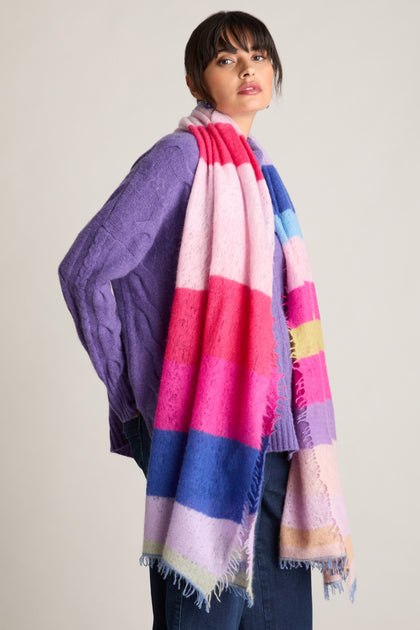 A person wearing a purple sweater, jeans, and a Rainbow Stripe Cashmere Scarf stands against a plain background, showcasing the perfect Autumn look.