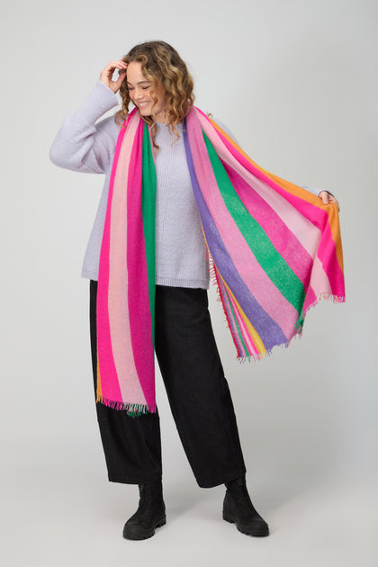 A woman smiles in a light sweater, dark pants, black boots, and holds the Colour Pop Stripe Cashmere Scarf featuring vibrant pink, purple, green, and orange stripes.