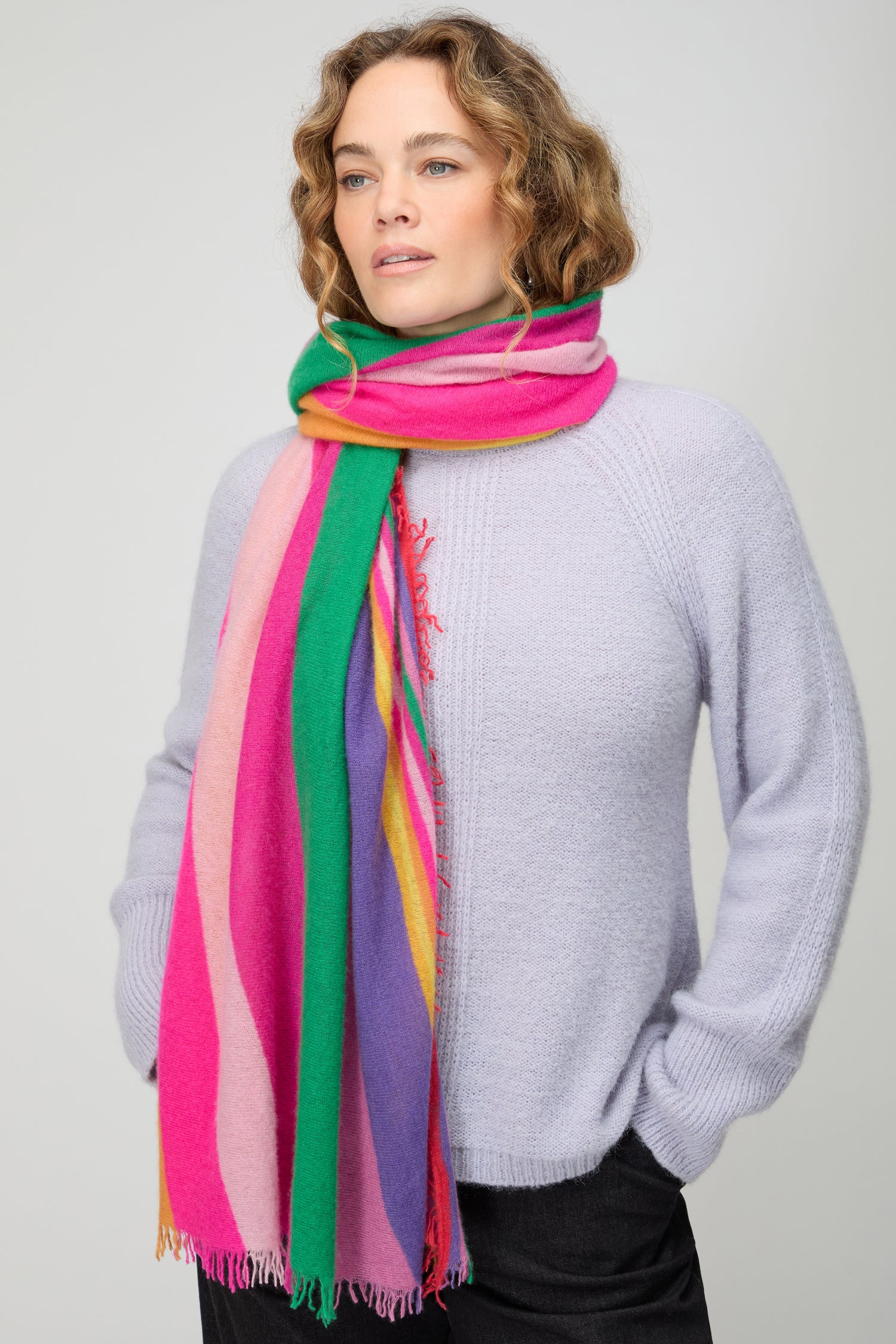 A person wearing a light purple sweater and the Colour Pop Stripe Cashmere Scarf stands with hands in pockets against a plain background.