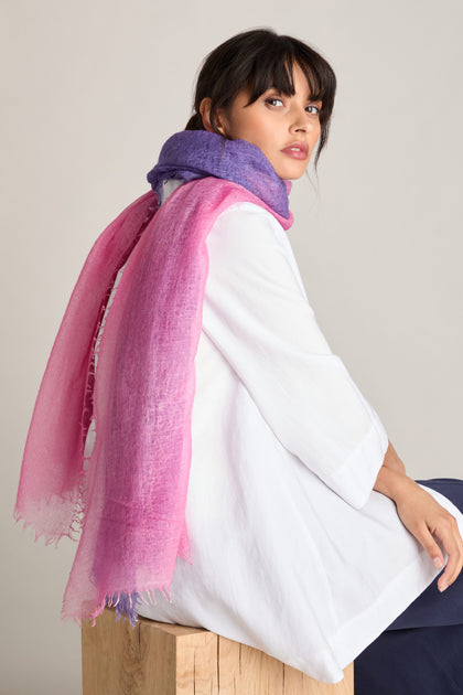 A woman wearing a white top and the Harmony Cashmere Scarf in pink lilac sits on a wooden stool against a plain background, making it a perfect Autumn accessory.