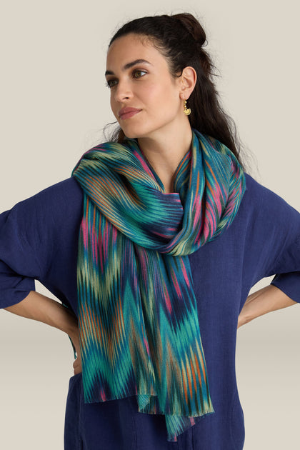 Person clad in an Autumnal Ikat Wool Scarf, featuring a zigzag pattern, layered over a blue top, standing with hands on hips and gazing to the side.