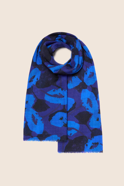 The Flowering Poppies Wool Scarf, featuring a luxurious blue and black pattern, is elegantly folded in a circular shape and displayed on a flat beige surface, making it the perfect addition to elevate your winter wardrobe.