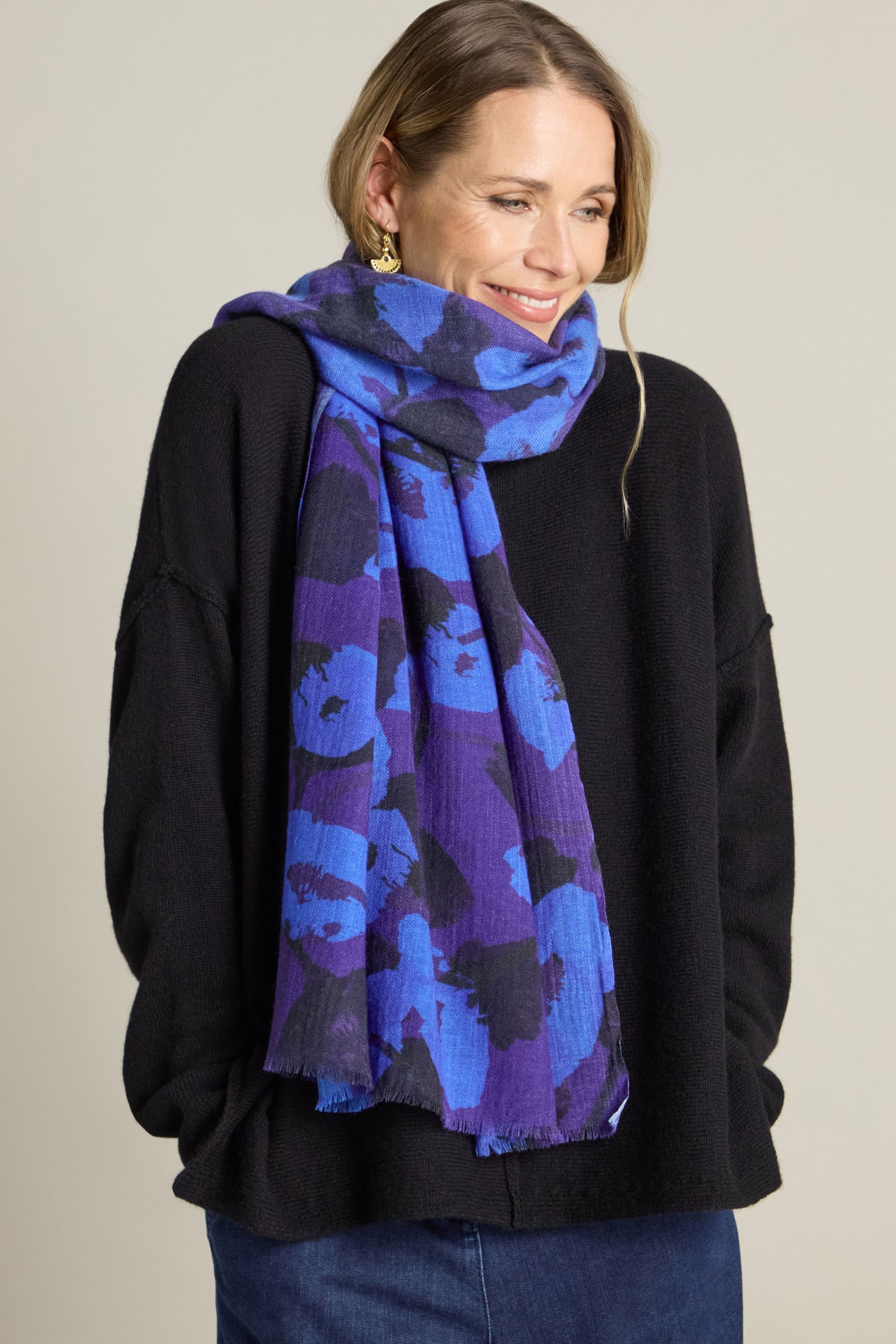 A person wearing a black sweater and a sumptuous Flowering Poppies Wool Scarf smiles with hands in pockets against a plain background, highlighting their winter wardrobe.