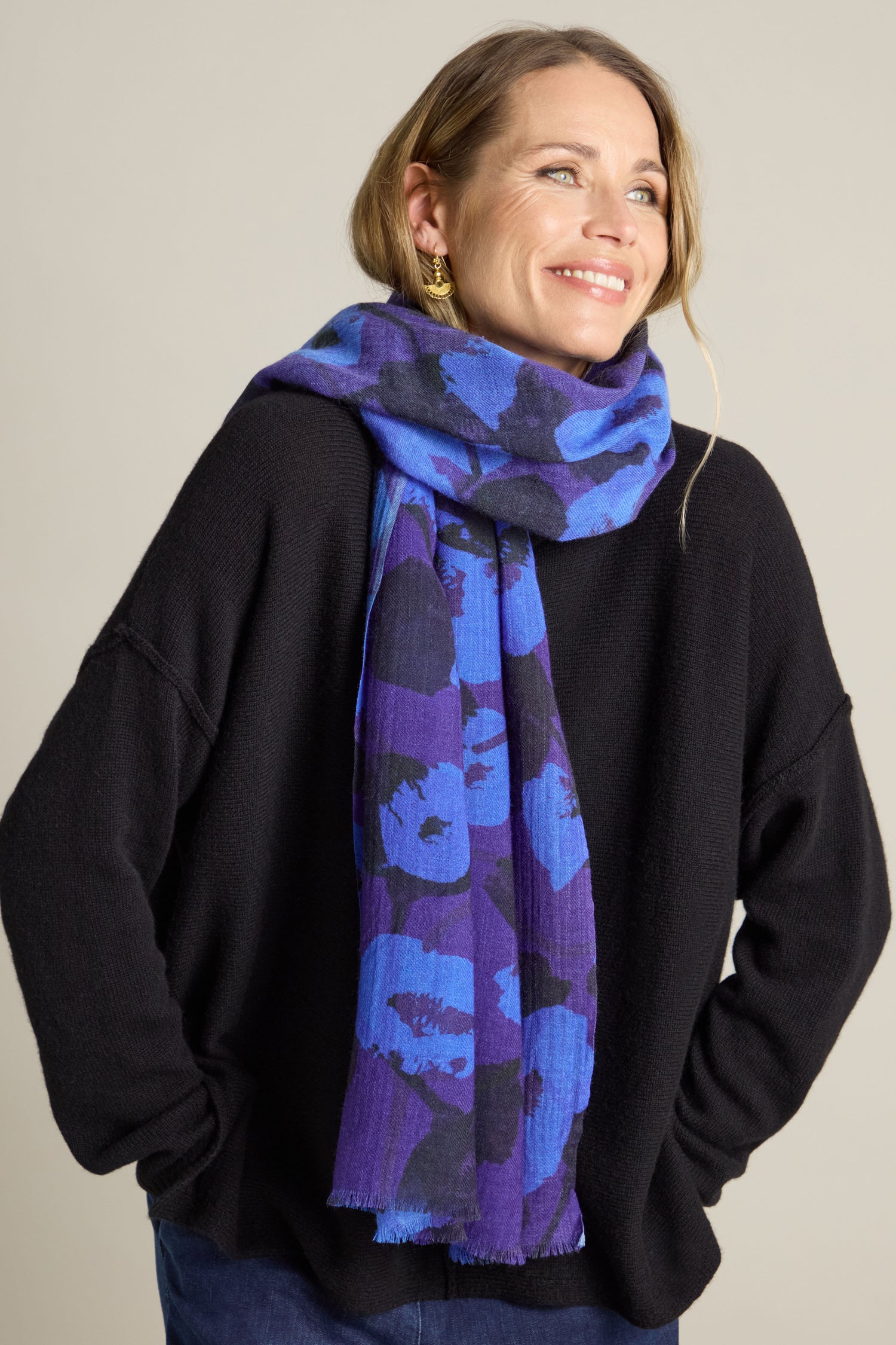 A person wearing a black sweater and the luxurious Flowering Poppies Wool Scarf, which features a stunning blue and purple pattern, smiles while looking slightly off camera.