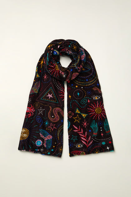 The Dream Stitched Print Scarf is a lightweight black scarf adorned with artisanal embroidery in colorful celestial and nature-inspired designs—stars, planets, and sea creatures—displayed flat on a light surface.