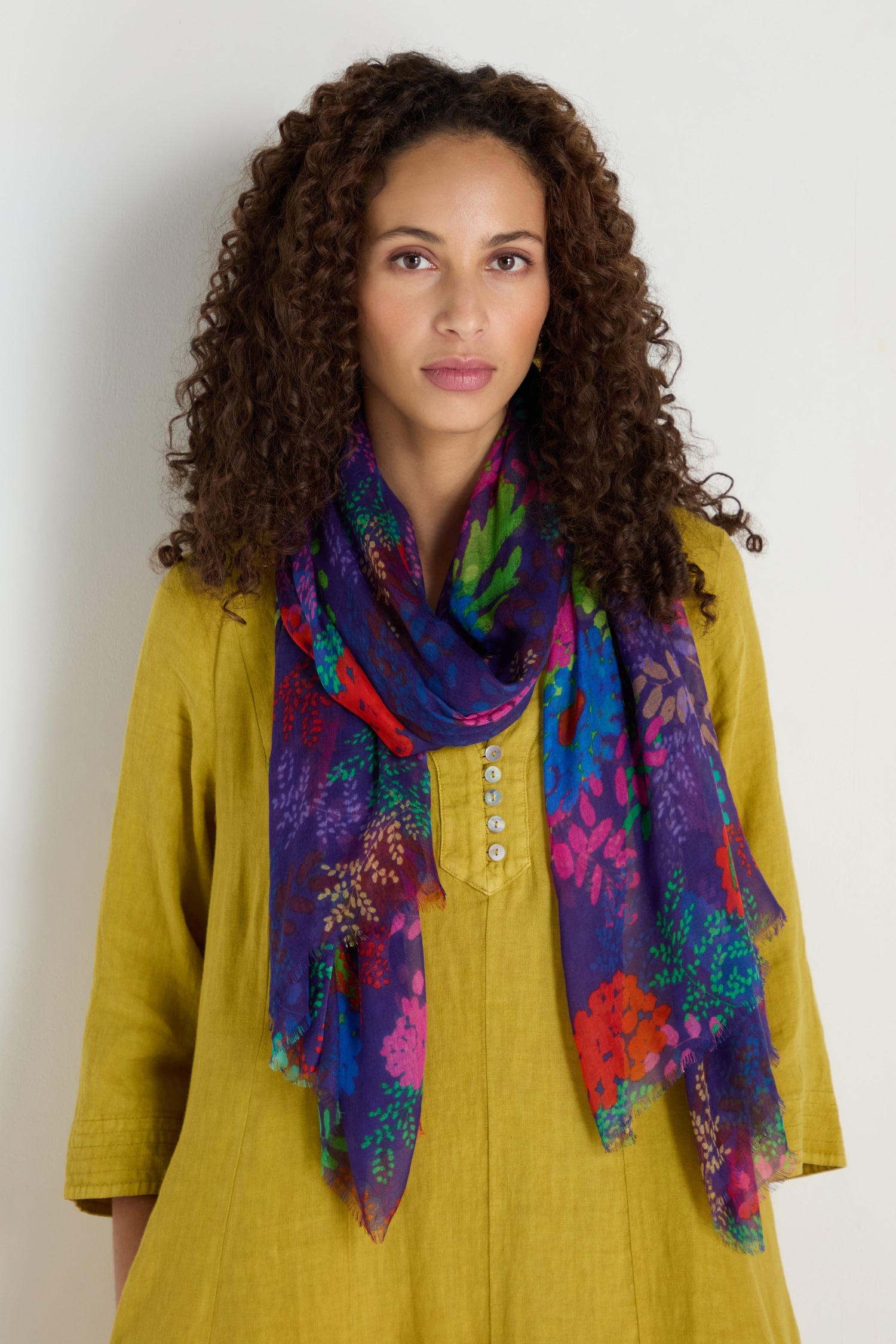 Woman with curly hair wearing the Bright Blooming Garden Scarf and a yellow buttoned dress, standing against a plain light-colored background.