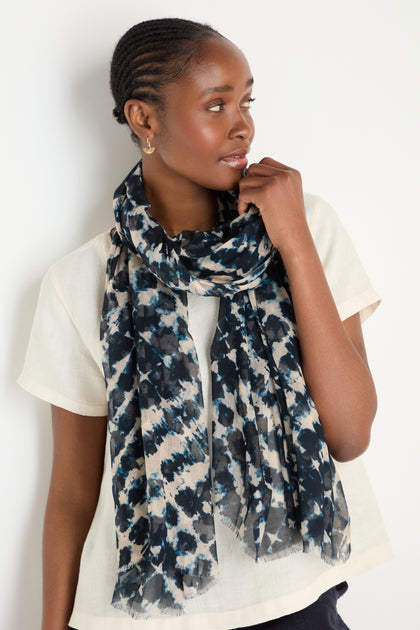 A woman wearing a short-sleeve cream blouse and the Diamond Tie Dye Scarf—featuring blue, black, and beige hues with a silky drape—looks to the side with one hand gently touching her face.