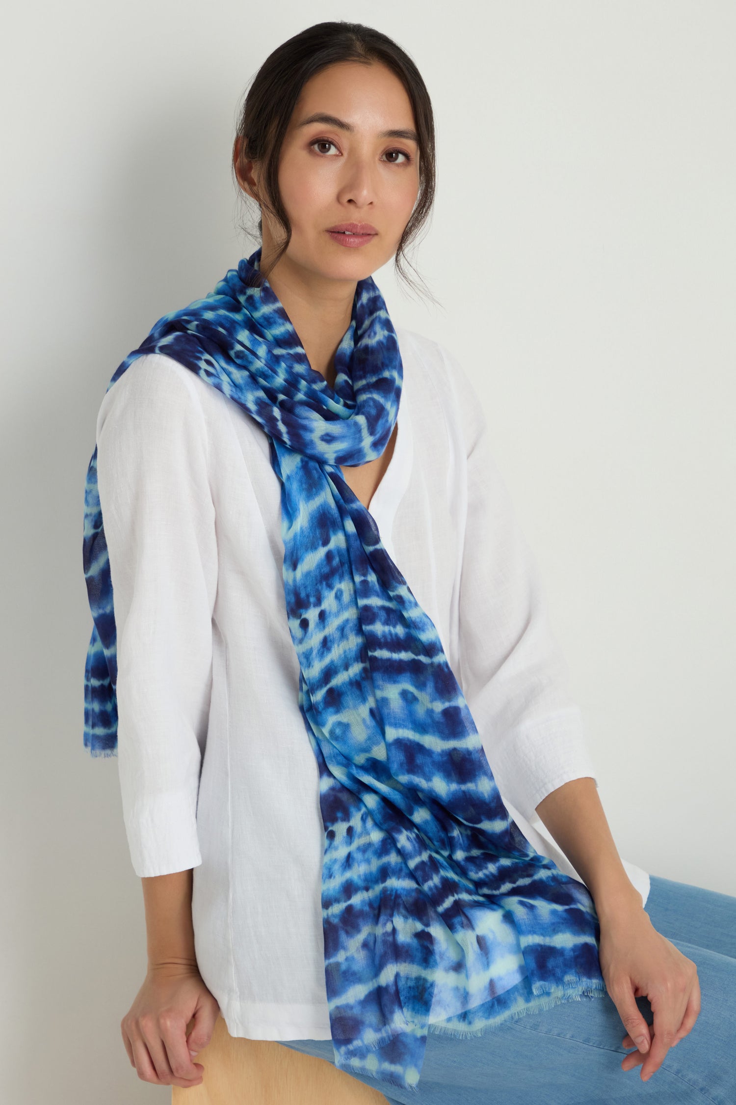 Woman wearing a white blouse and a blue tie dye print scarf sits on a wooden chair against a plain light background.