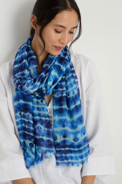 A woman in a white top wears the Tie Dye Print Scarf in blue modal silk, looking down with a neutral expression against a plain background.