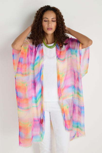 A woman stands against a plain background, eyes closed, hands raised to her head, wearing a colorful Painted Horizon Kimono over a white outfit.