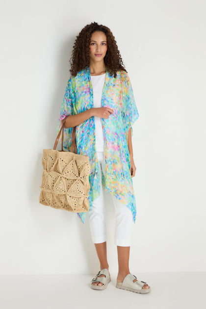 A woman stands against a white wall wearing white pants, a white top, the Summer Confetti Burst Kimono in vibrant patterns, beige sandals, and carrying a large woven tote bag.