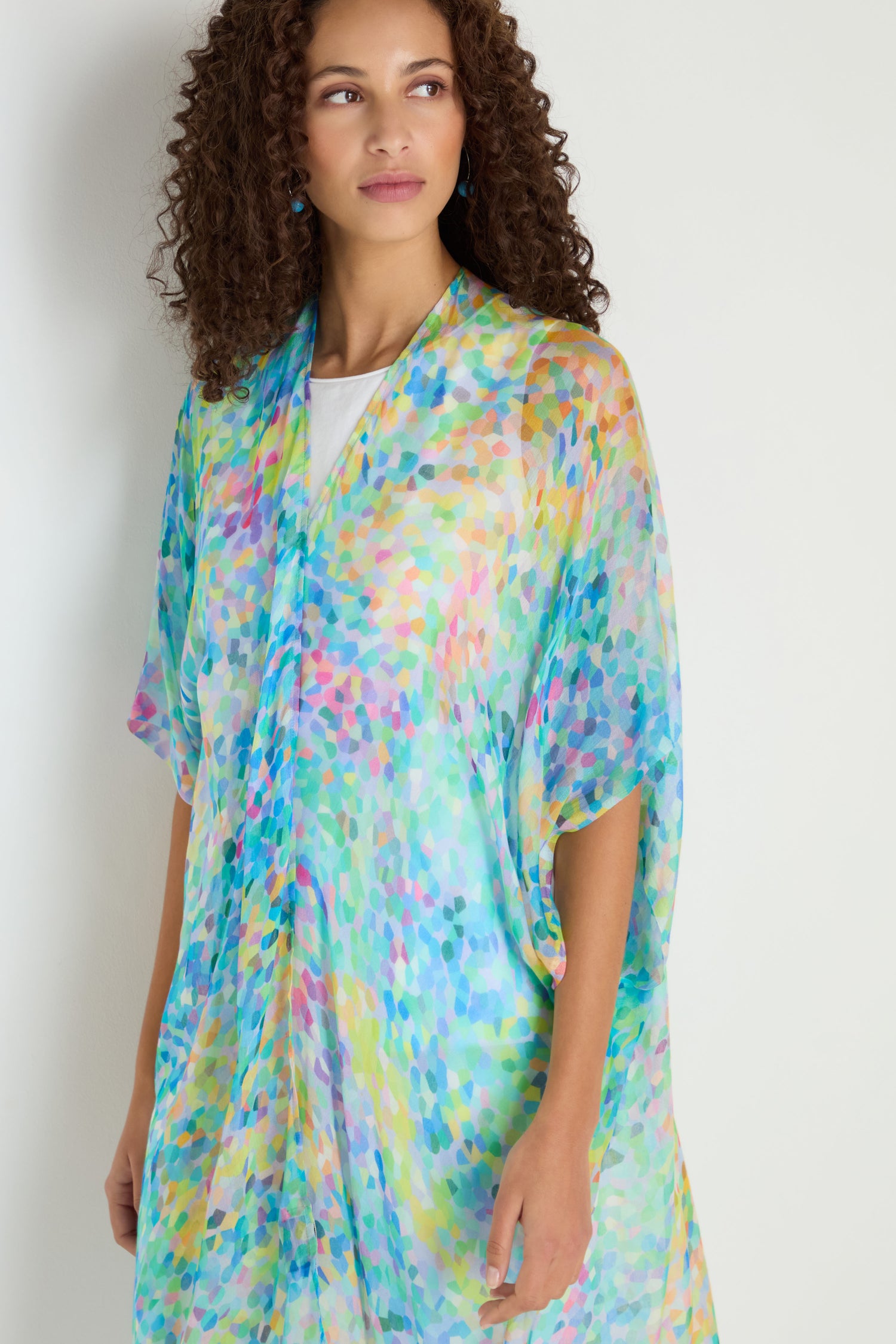 A woman with curly hair wears the Summer Confetti Burst Kimono, a loose, colorful, semi-transparent layer with a multicolored abstract pattern over a white top—an ideal lightweight cover-up.