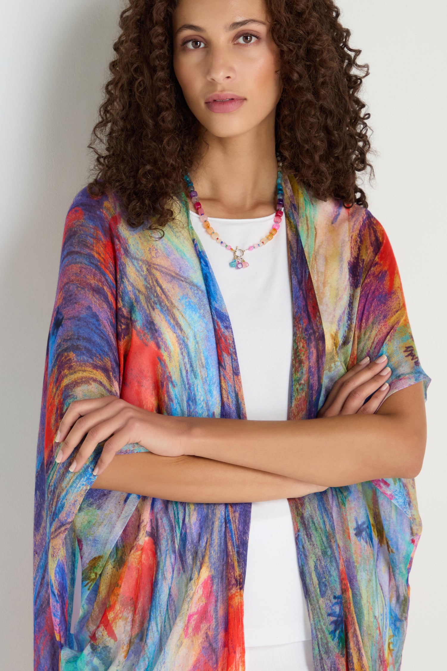 A woman with curly hair stands with arms crossed, wearing the Soft Meadow Dreams Kimono over a white top and accessorized with a beaded necklace.
