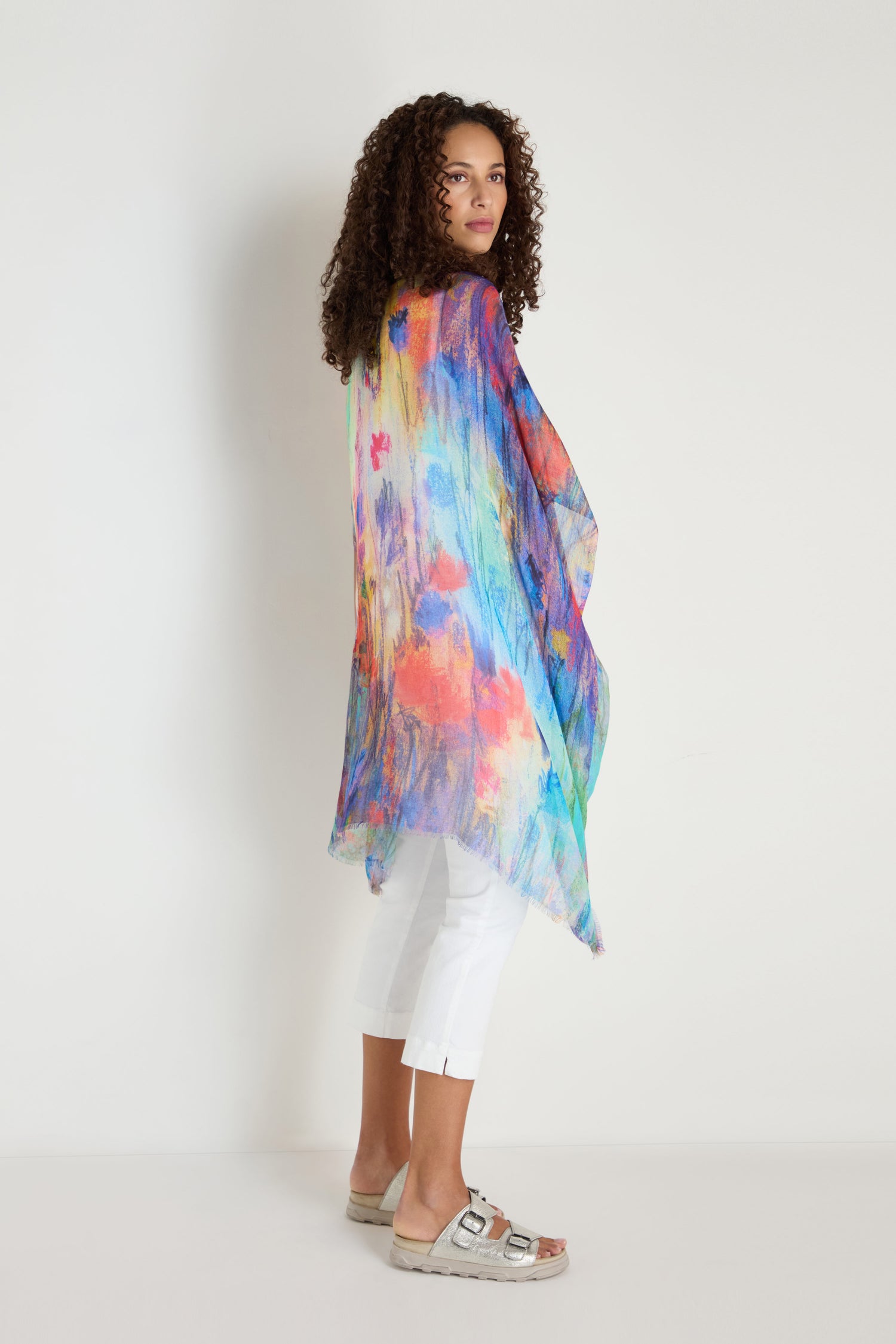 A woman stands sideways against a white wall, wearing the Soft Meadow Dreams Kimono with white pants and beige sandals.